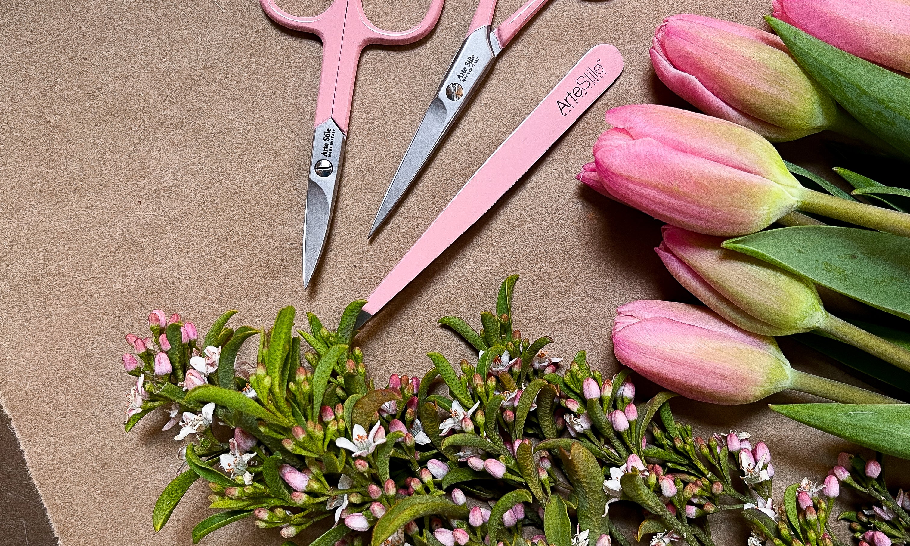 ArteStile Brow Scissors, ArteStile Nail Scissors, and ArteStile tweezers in rosé (light pink), on a brown wood table with pink tulips. 