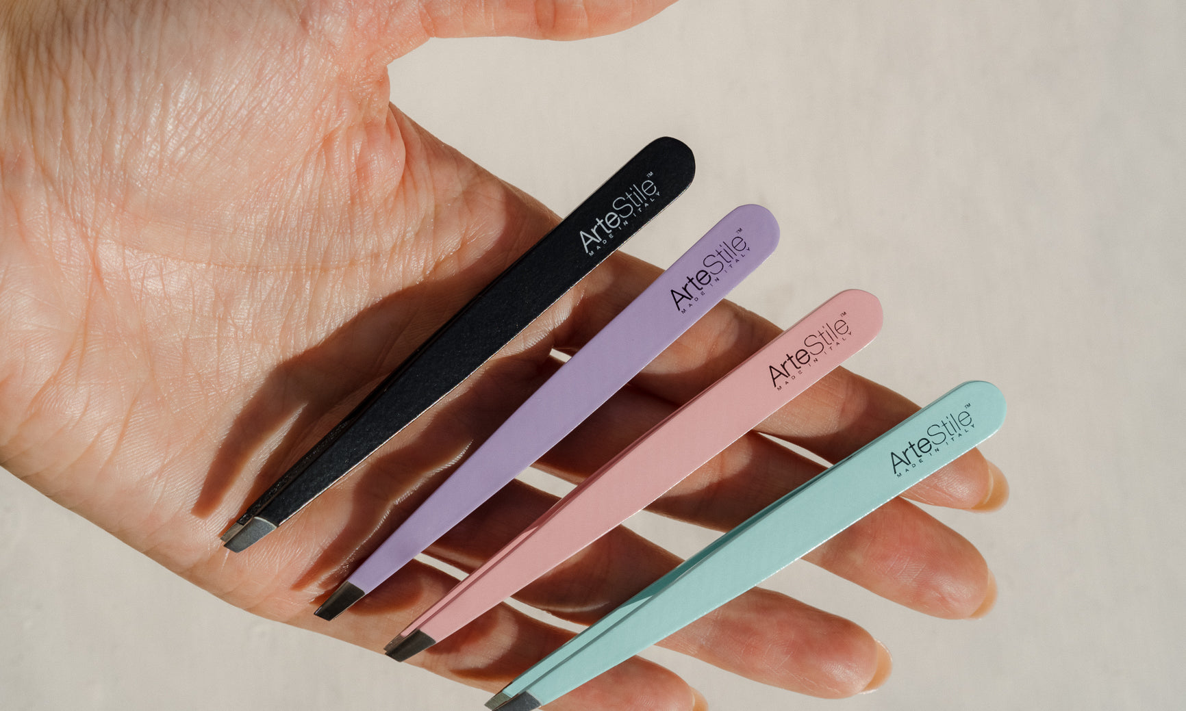 A hand holding four pairs of tweezers. One is black, one is purple, one is pink, and one is mint.